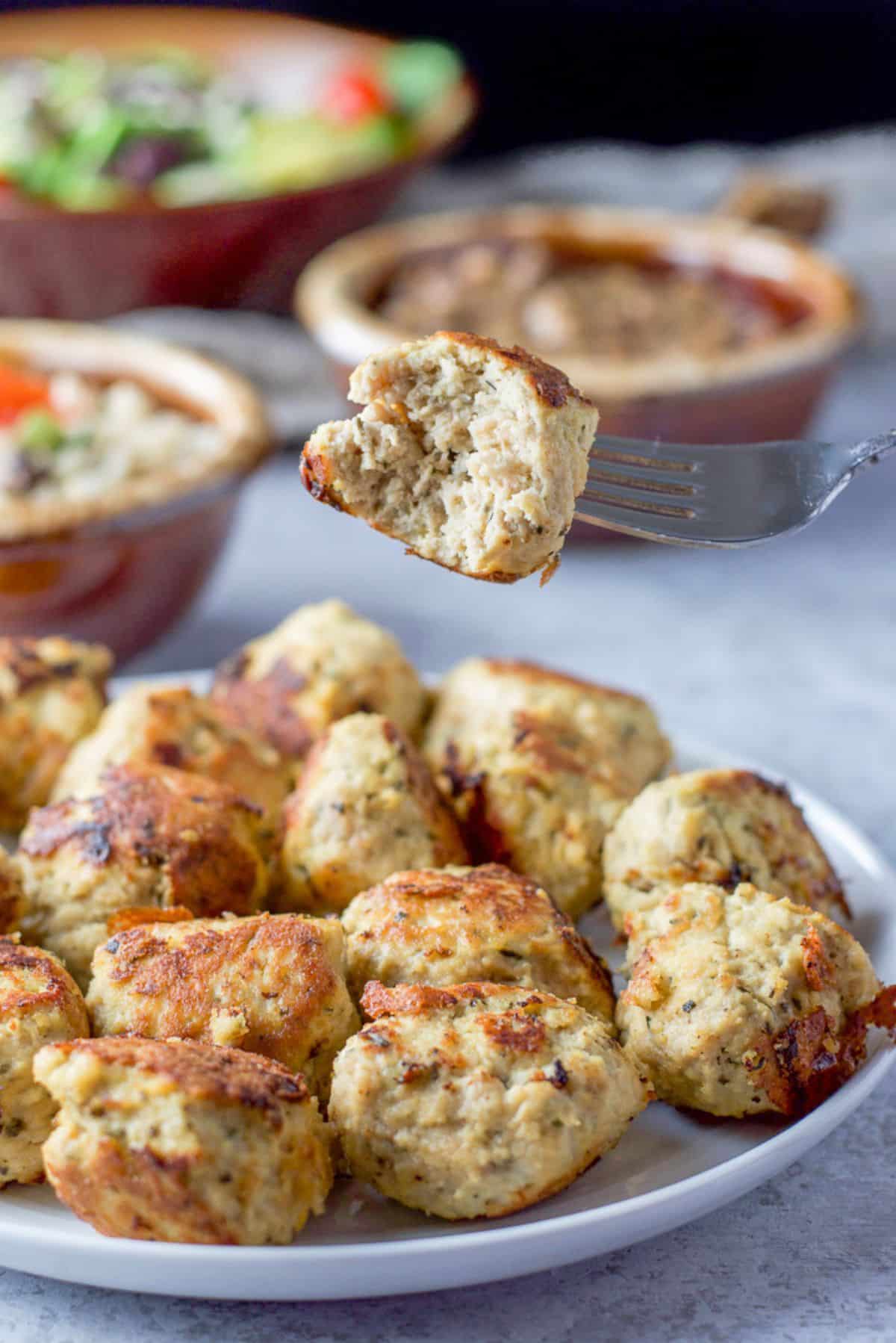 a fork with a bite taken out of it held over the plate of meatballs with other dishes in back