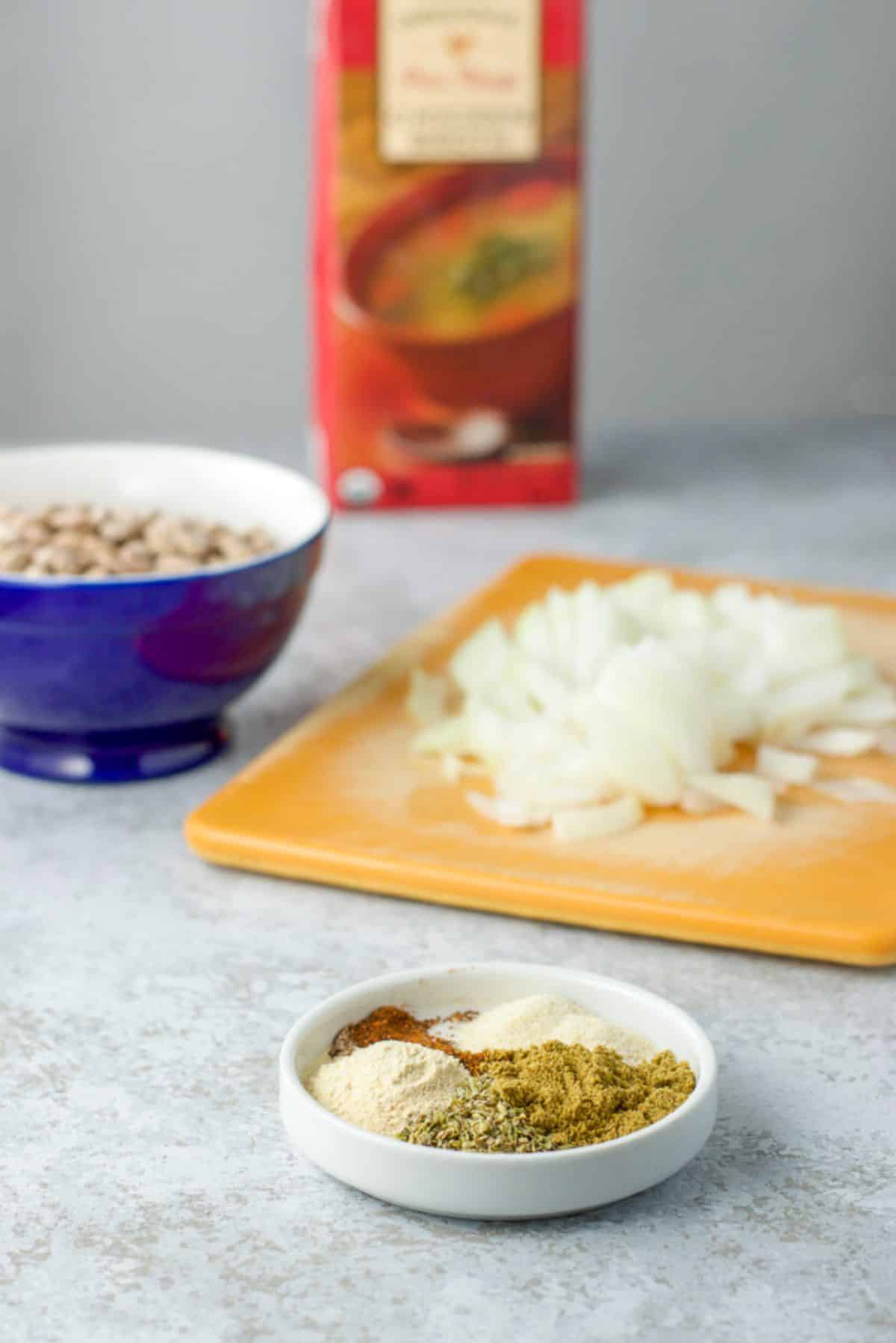 Oregano, cumin, garlic powder, onion powder and chili powder on a dish, with onions chopped, beans in a bowl and broth