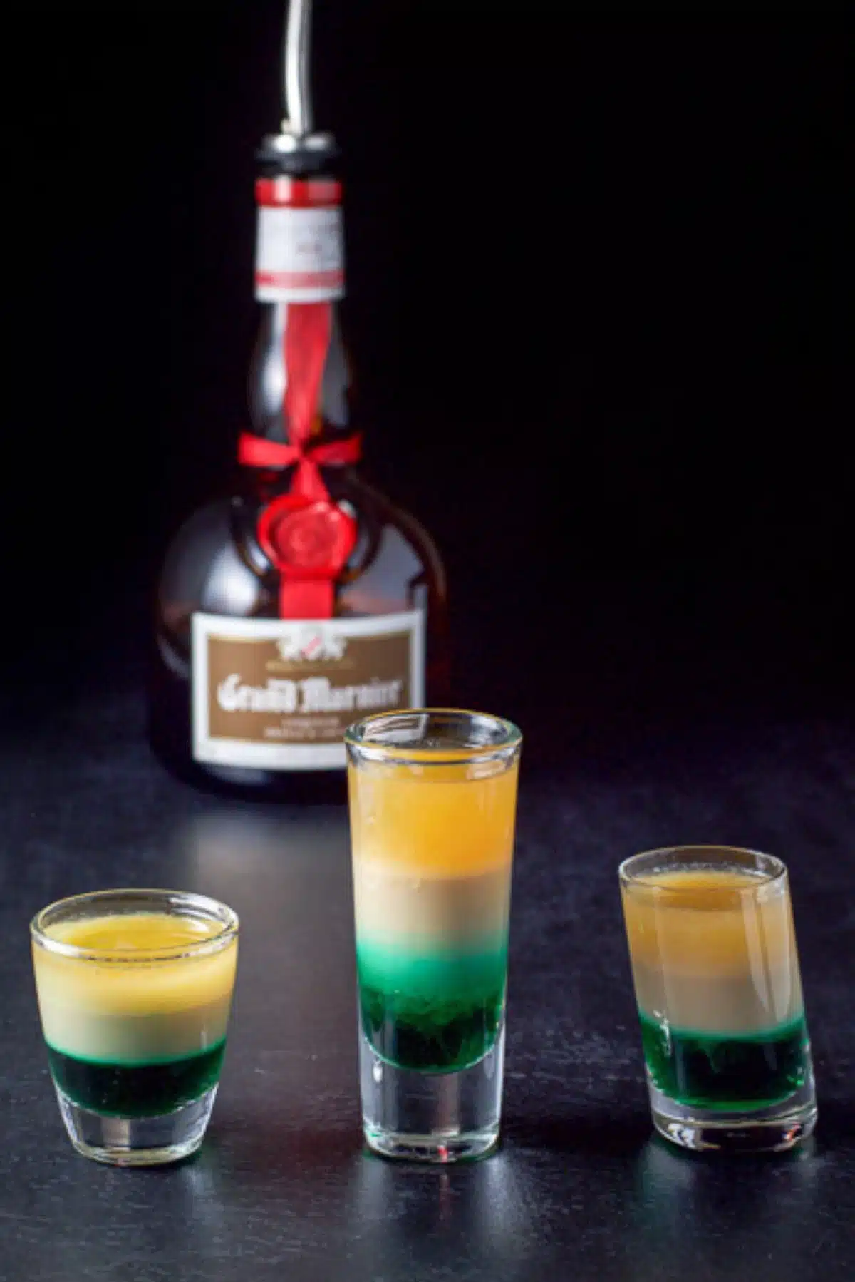 The three glasses filled with the green, beige and yellow shots in front of the bottle of grand marnier