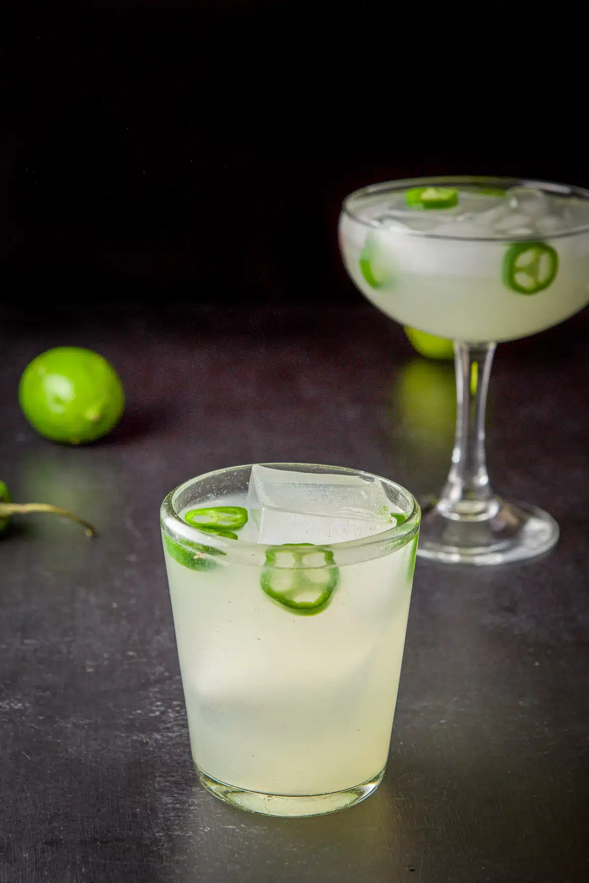 Two margarita glasses on a table with the jalapeno margarita in it along with slices