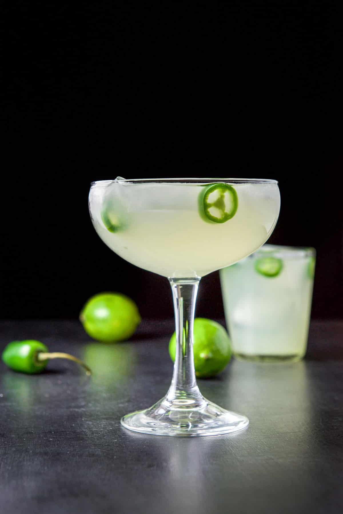 Vertical view of the two margarita glasses filled with the cocktail with jalapeno slices in it and limes on the table