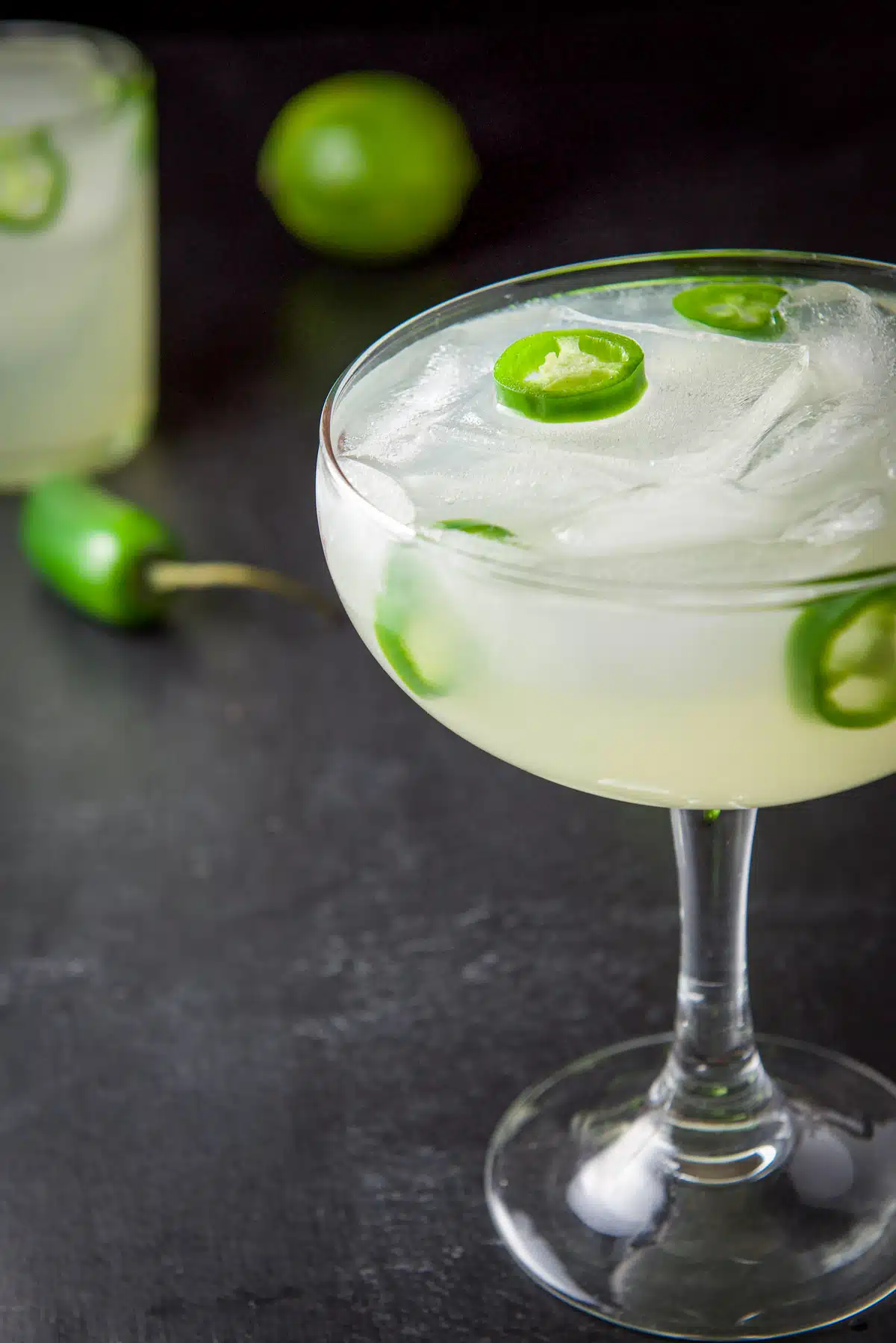A bowl margarita glass off to the side filled with the margarita with jalapeno slices on it