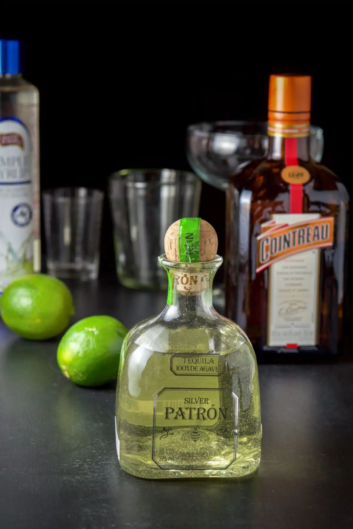 Tequila, limes, orange liqueur, and simple syrup on a table with glassware