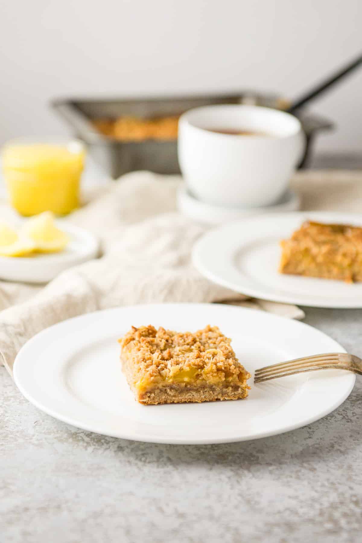 Lemon bars on two white plates with tea and the pan of bars in the background