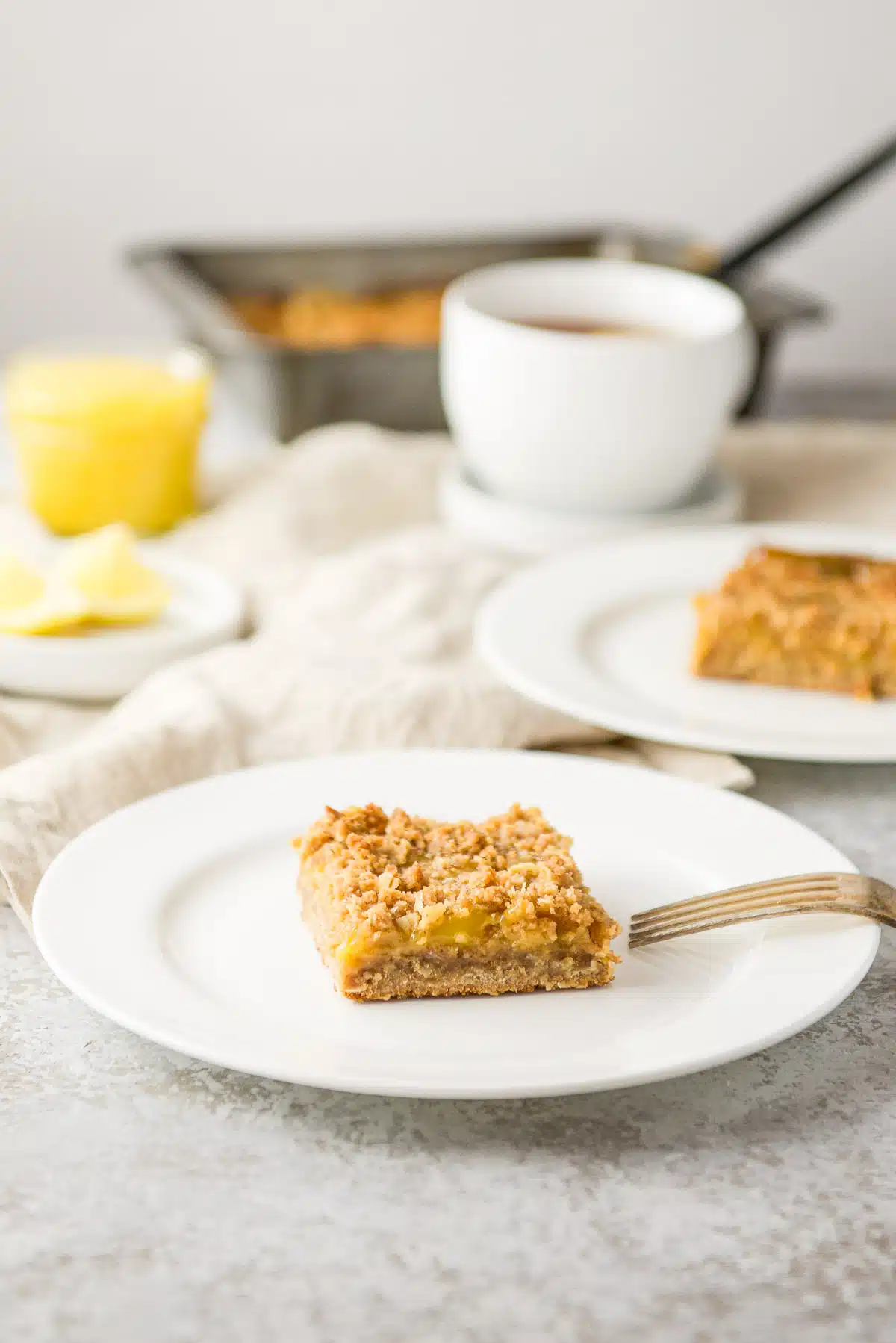 Lemon bars on two white plates with tea and the pan of bars in the background