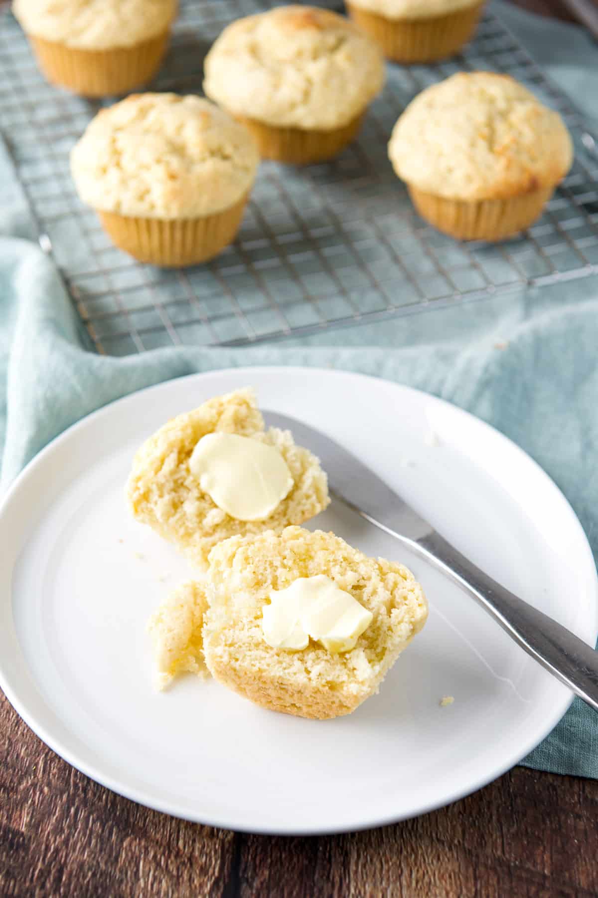 A white plate with the two halves of the lemon muffin with butter on it. There is also a rack in the background with muffins on it