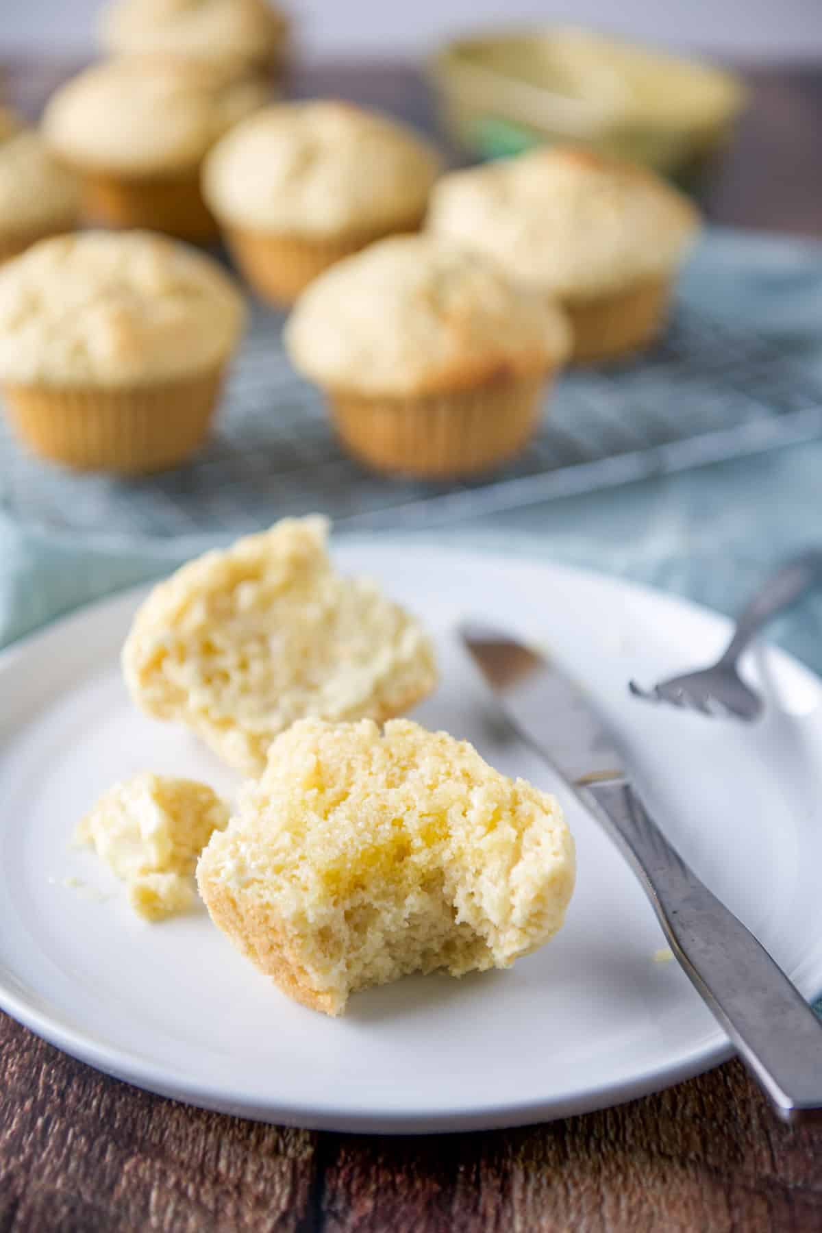 A bite taken out of the lemon muffin half with the rest of the muffin on the plate and more muffins in back