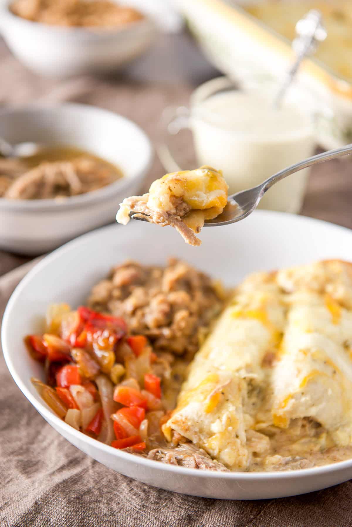 A forkful of enchilada with pork in it held over a plate with enchiladas, beans and veggie. There are bowls of pulled pork, sauce and beans in the background
