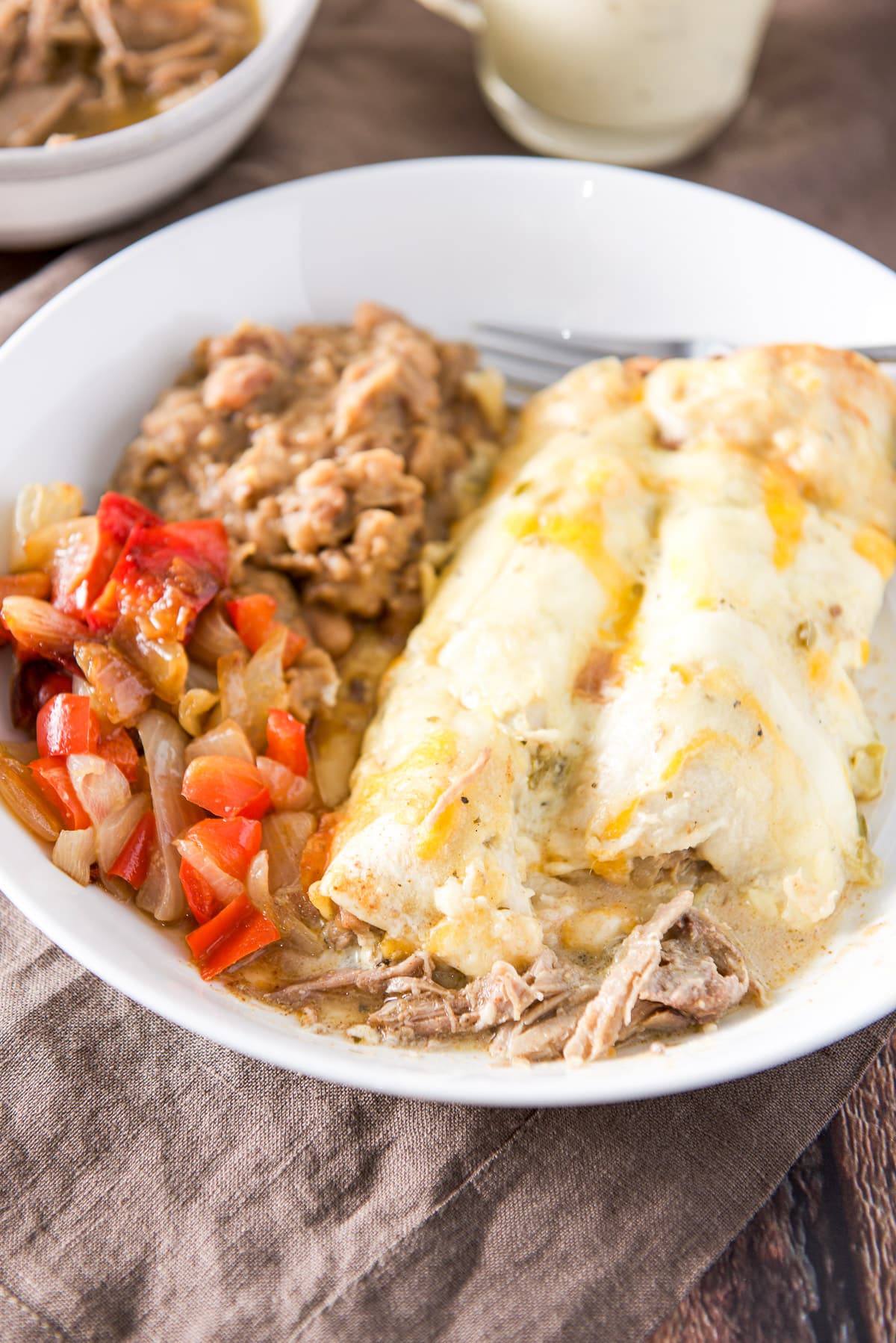 Two enchiladas with sour cream verde sauce with pork hanging out of them. There is also vegetables and beans on the plate