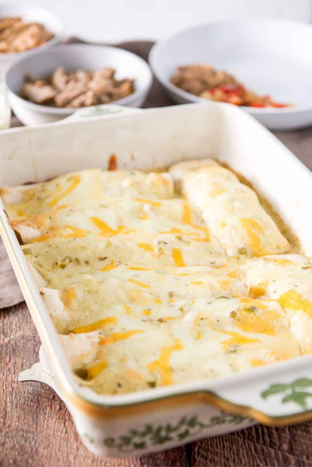 The enchiladas with sour cream sauce on the enchiladas in a baking dish and straight out of the oven. There are beans and pork in the background along with a plate