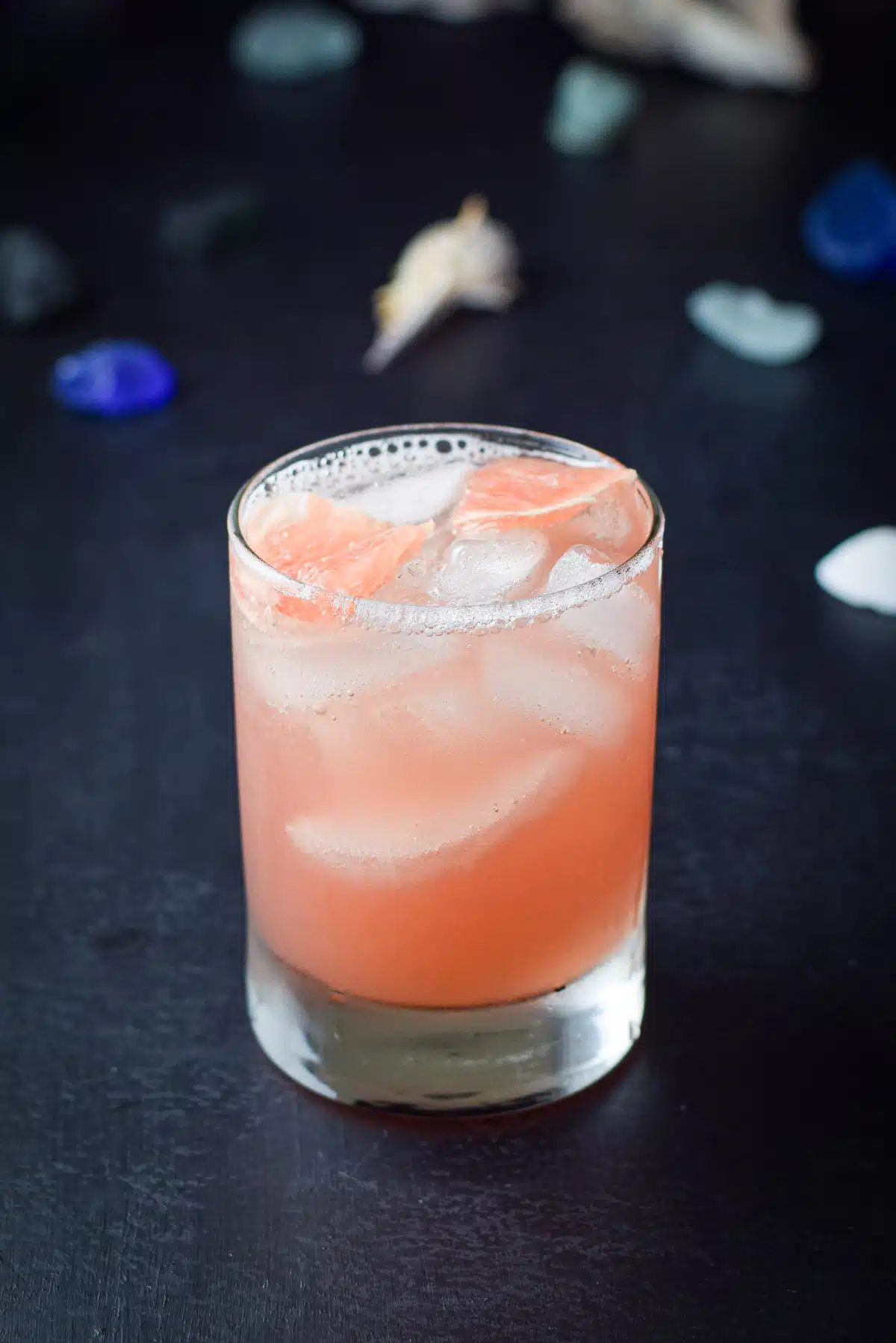 A old fashioned glass of the cocktail with grapefruit chunks in the drink and sea glass and shells in the back