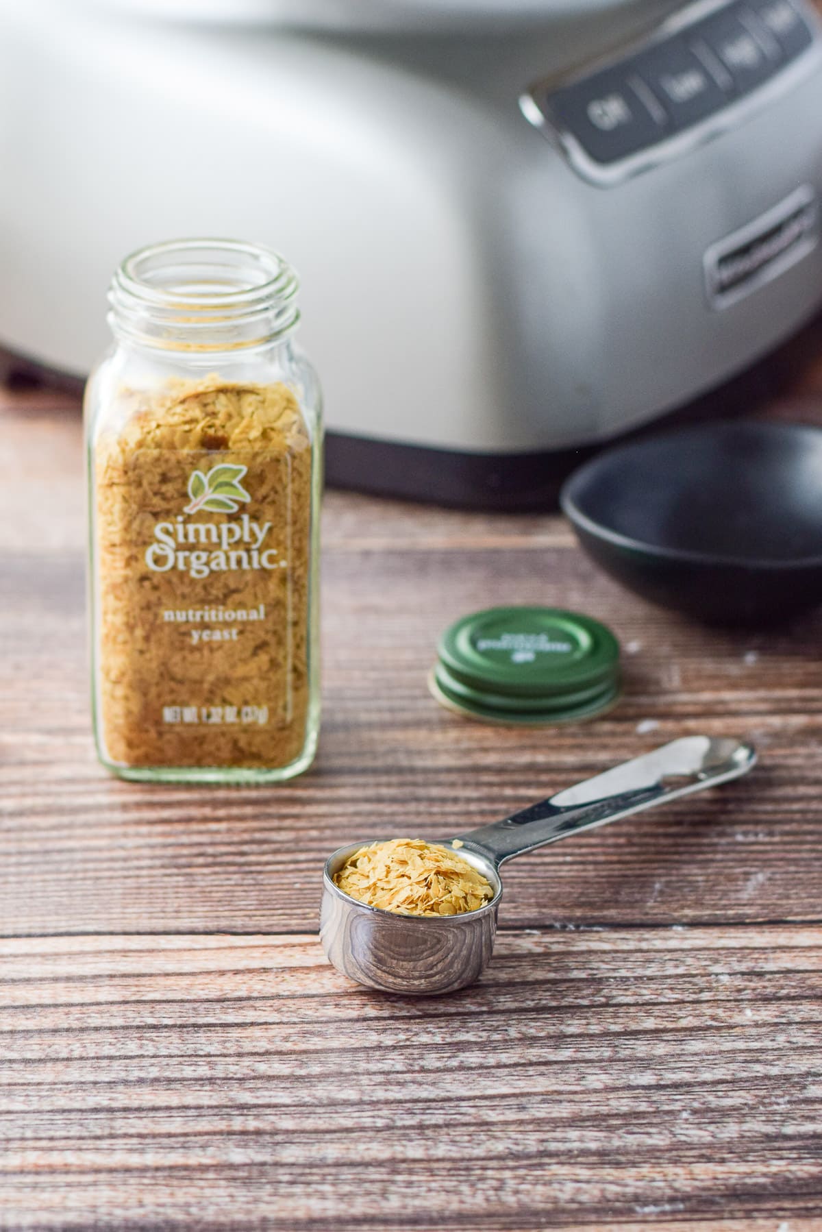 A tablespoon with nutrutional yeast in it in front of the processor and bottle