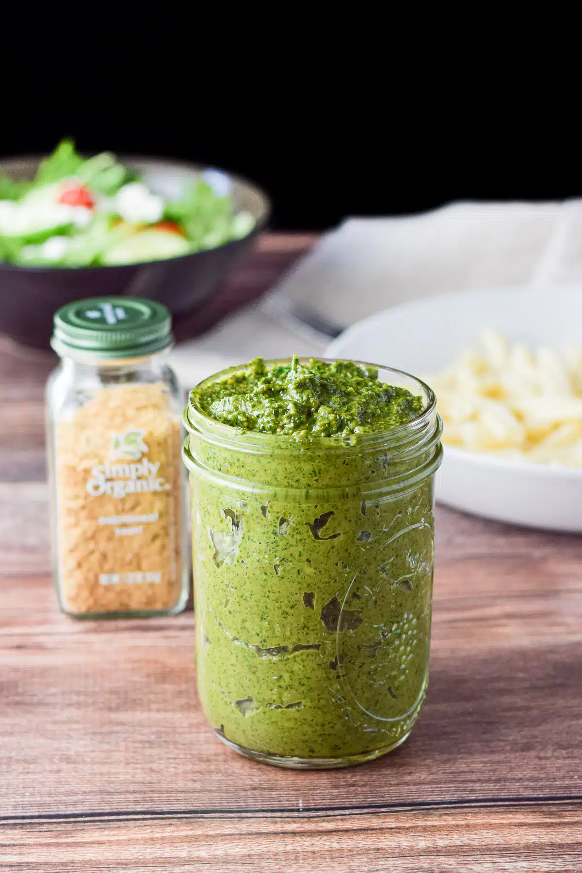 A jar with pesto in it, with nutritional yeast, pasta, and salad in the back