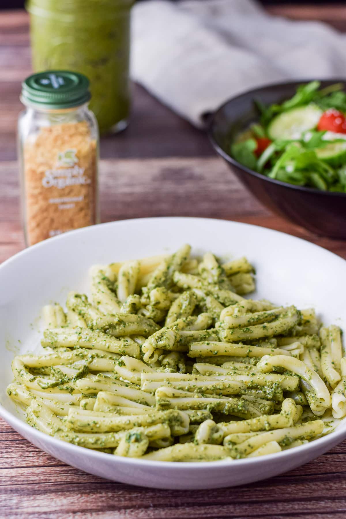A white dish with pasta with peston on it, with a salad in the back