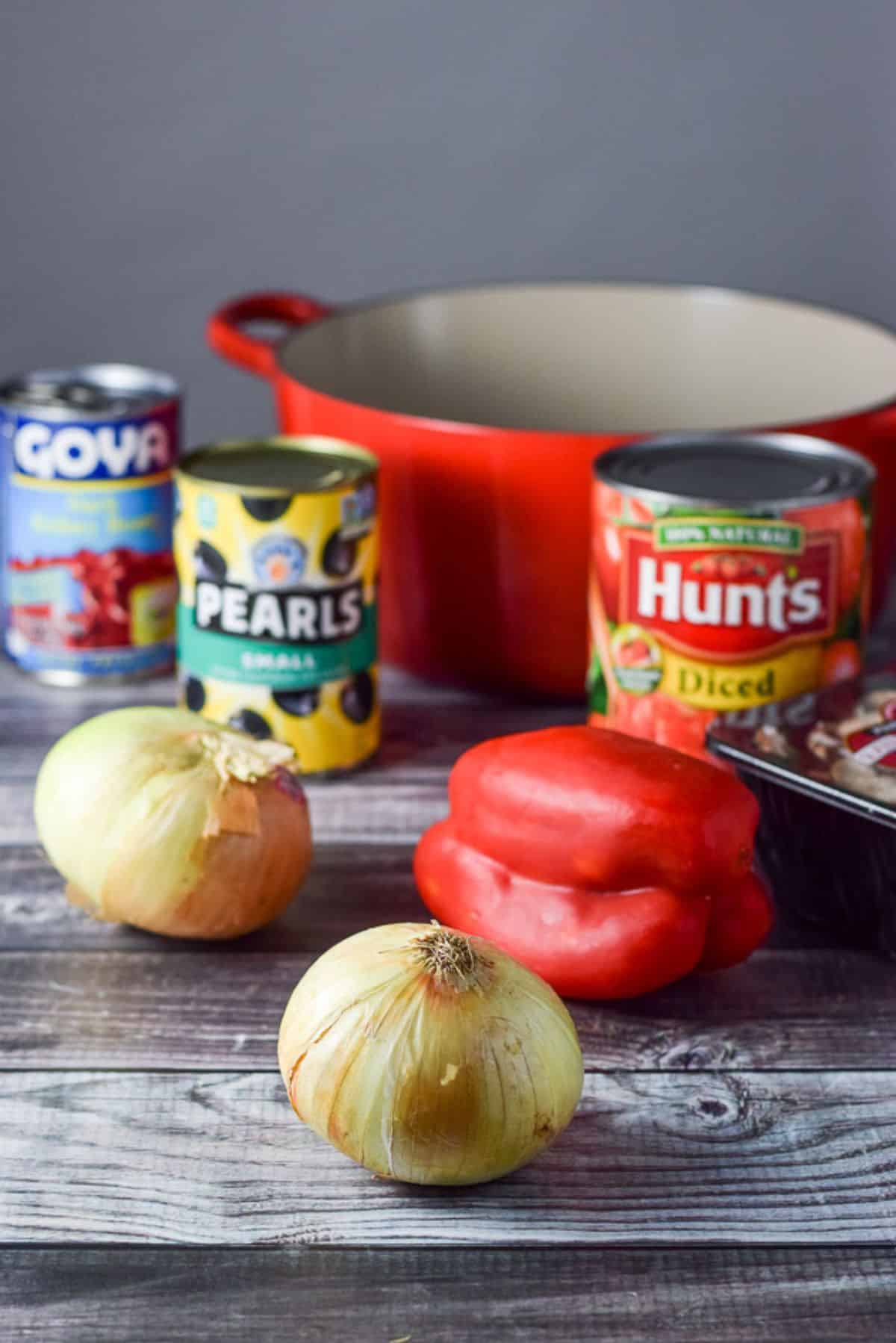 Onions, red bell pepper, mushrooms, diced tomatoes, black olives and red kidney beans on the table with a red Dutch oven