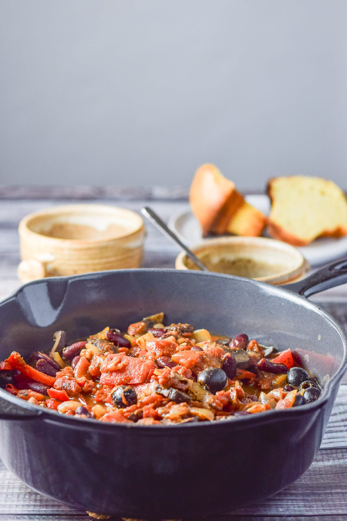 Pan of chili in a deep cast iron pan with two crocks and a corn muffin in the background