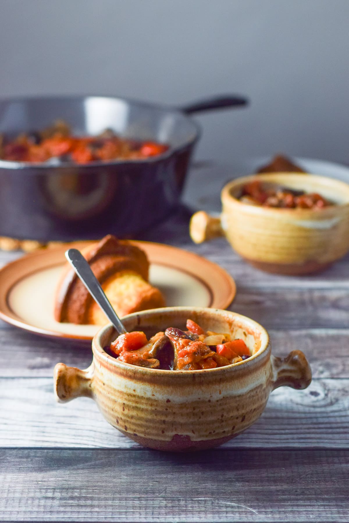 A crock filled with chili with a spoon in it and a corn muffin behind it and the pan and another crock
