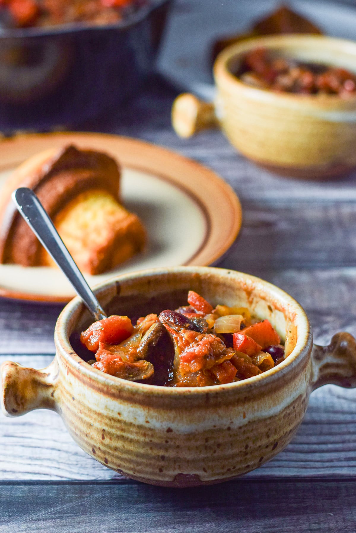 Close up of a crock of chili with a half of a corn muffin, another crock and pan of chili in the background