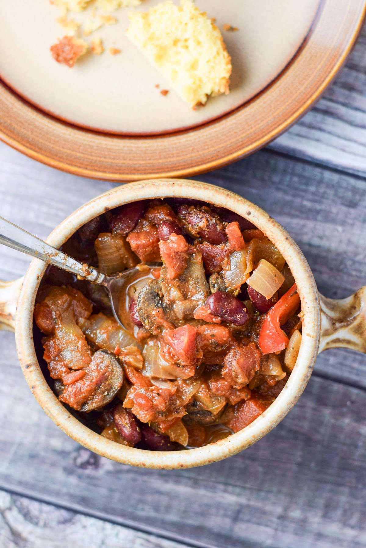 Overhead shot of a crock vegetarian chili with a spoon in it and a crumbled corn muffin in the background