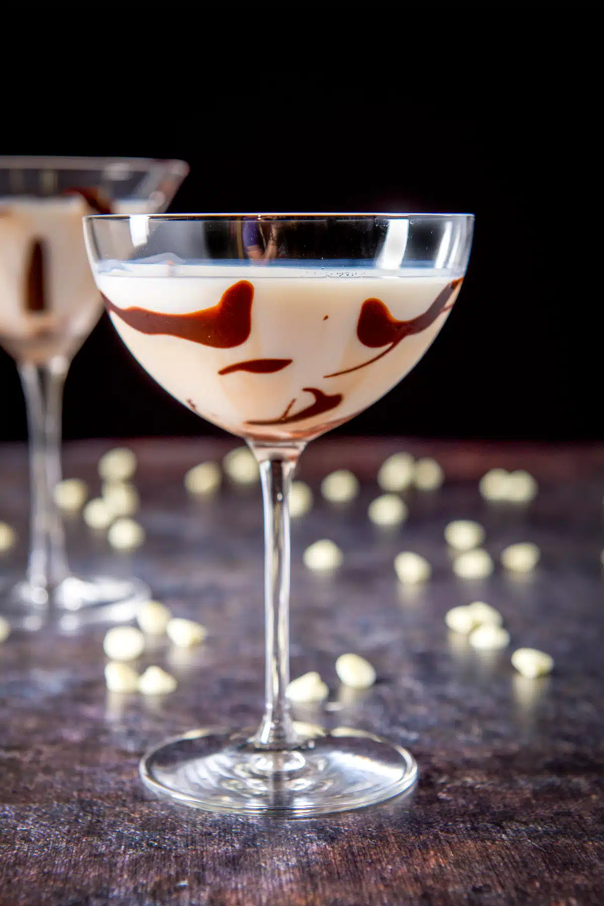 A bowl type of martini glass filled with the white chocolate martini with chocolate chips on the table along with another glass