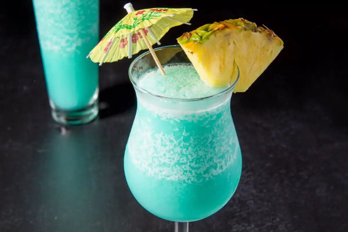 A tulip glass filled with a frozen blue drink with a pineapple wedge on the rim and a umbrella in the drink - square