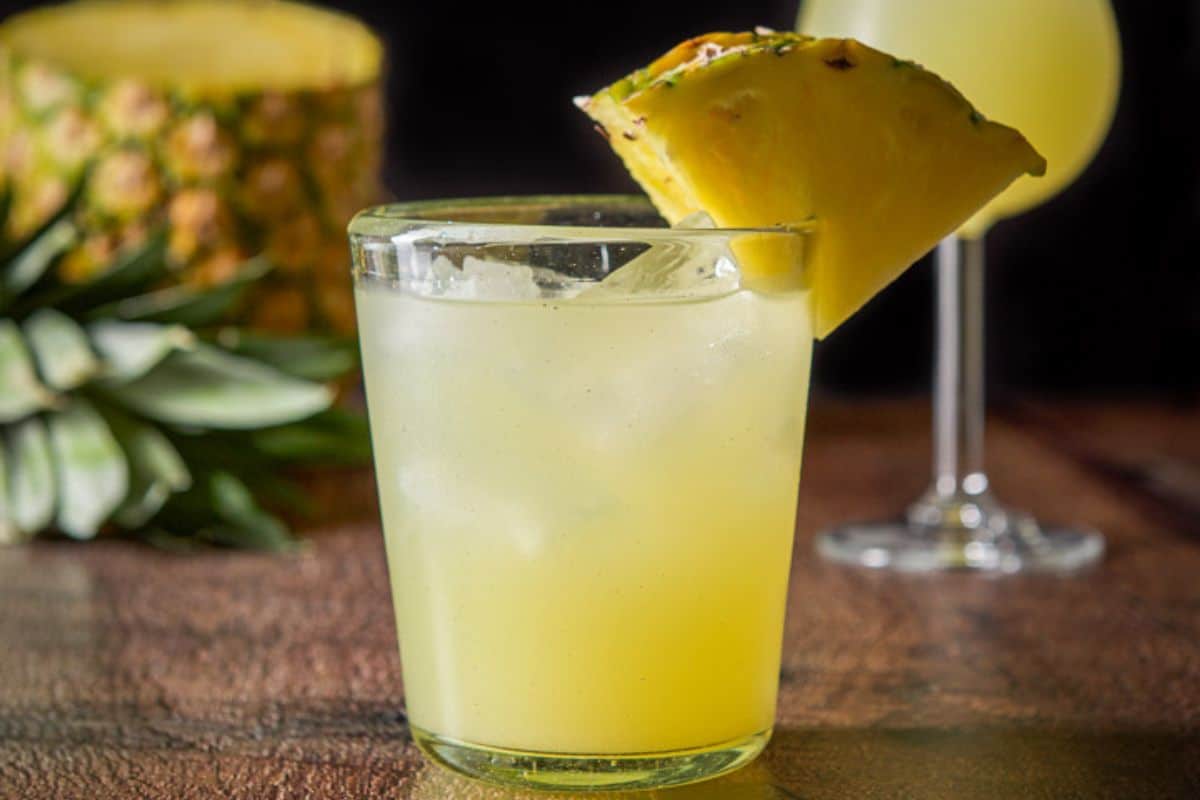 Double old fashioned glass filled with the matador with two pineapples on the side of the glass. There is a tulip glass with the cocktail and a pineapple in the background - square