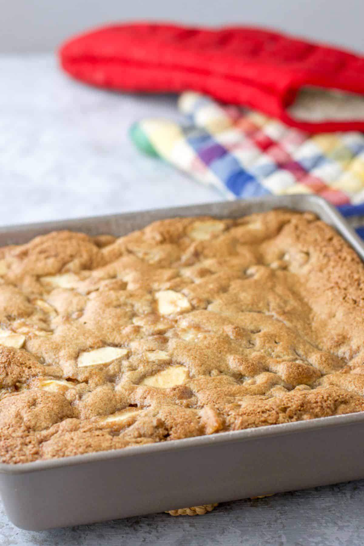 A square pan of apple cake with a few oven mitts in the background