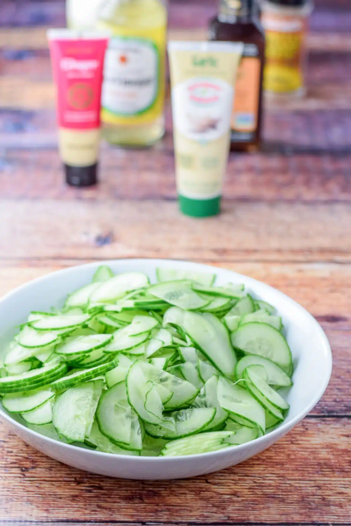 English cucumber sliced real thin and in a white bowl with dressing ingredients in the background
