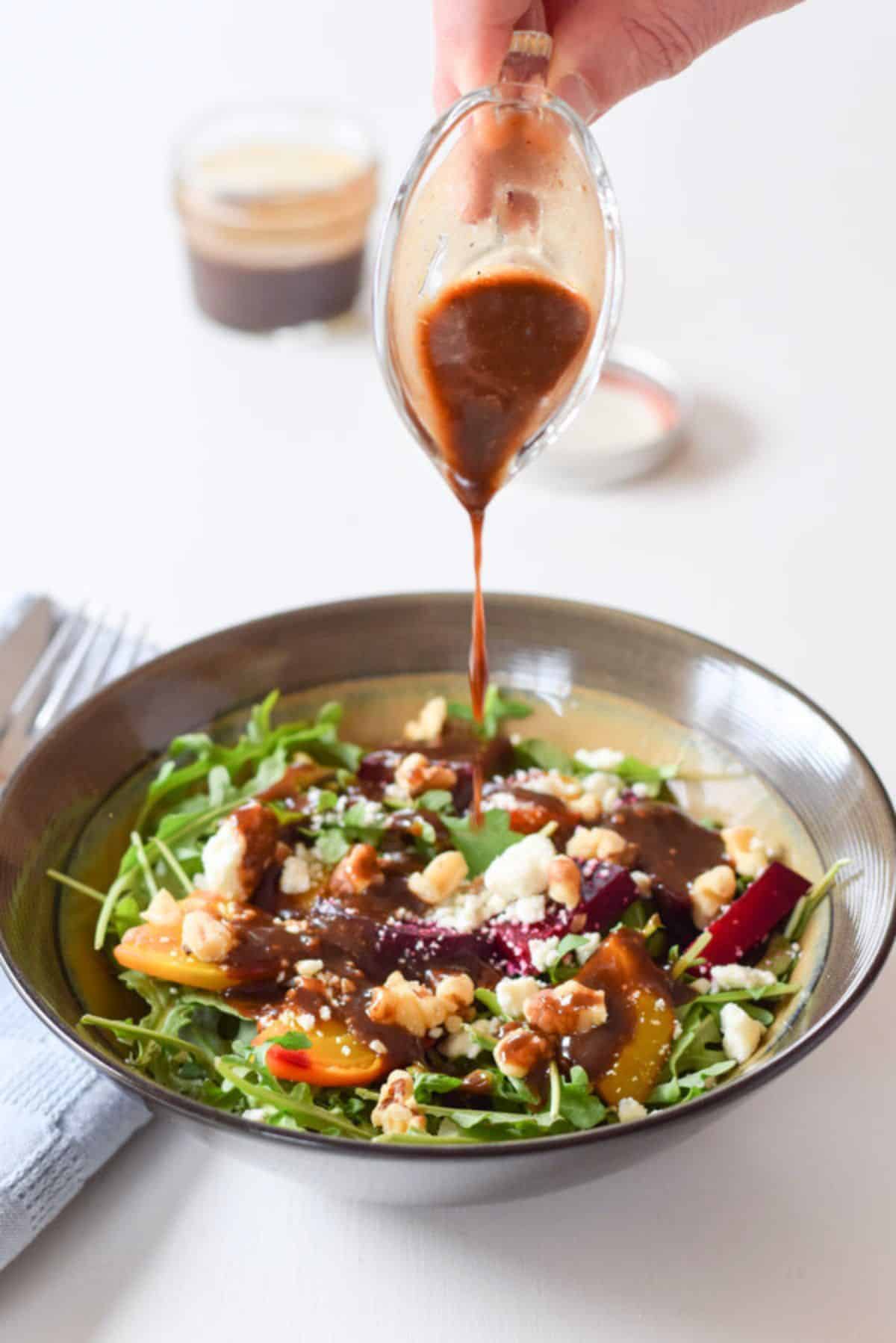 A hand holding a small glass gravy boat pouring the dressing on the salad