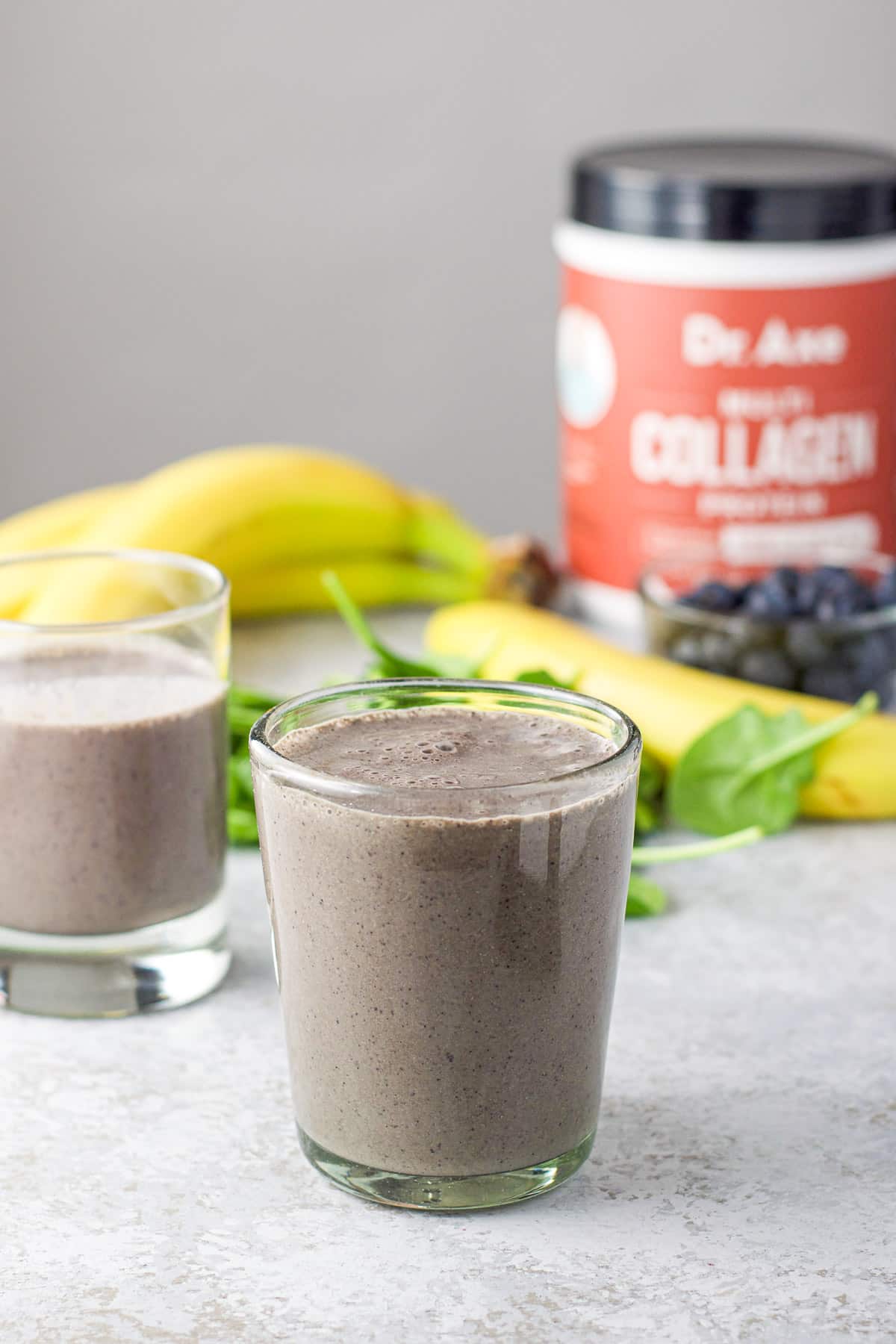 Two glasses filled with the spinach smoothie with collagen in the background as well as some blueberries and bananas