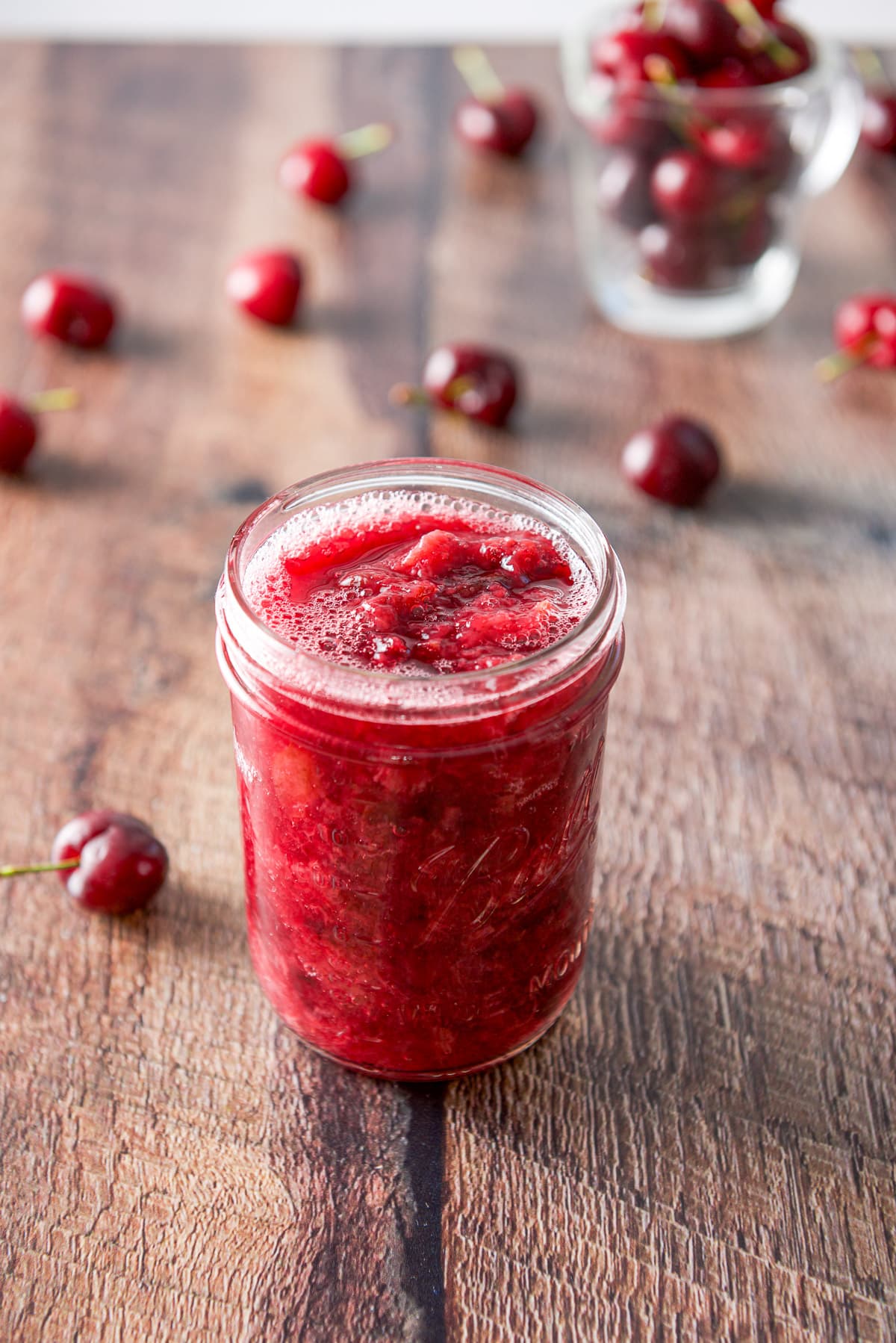 A jar of cherry sauce on the table with cherries strewn across it