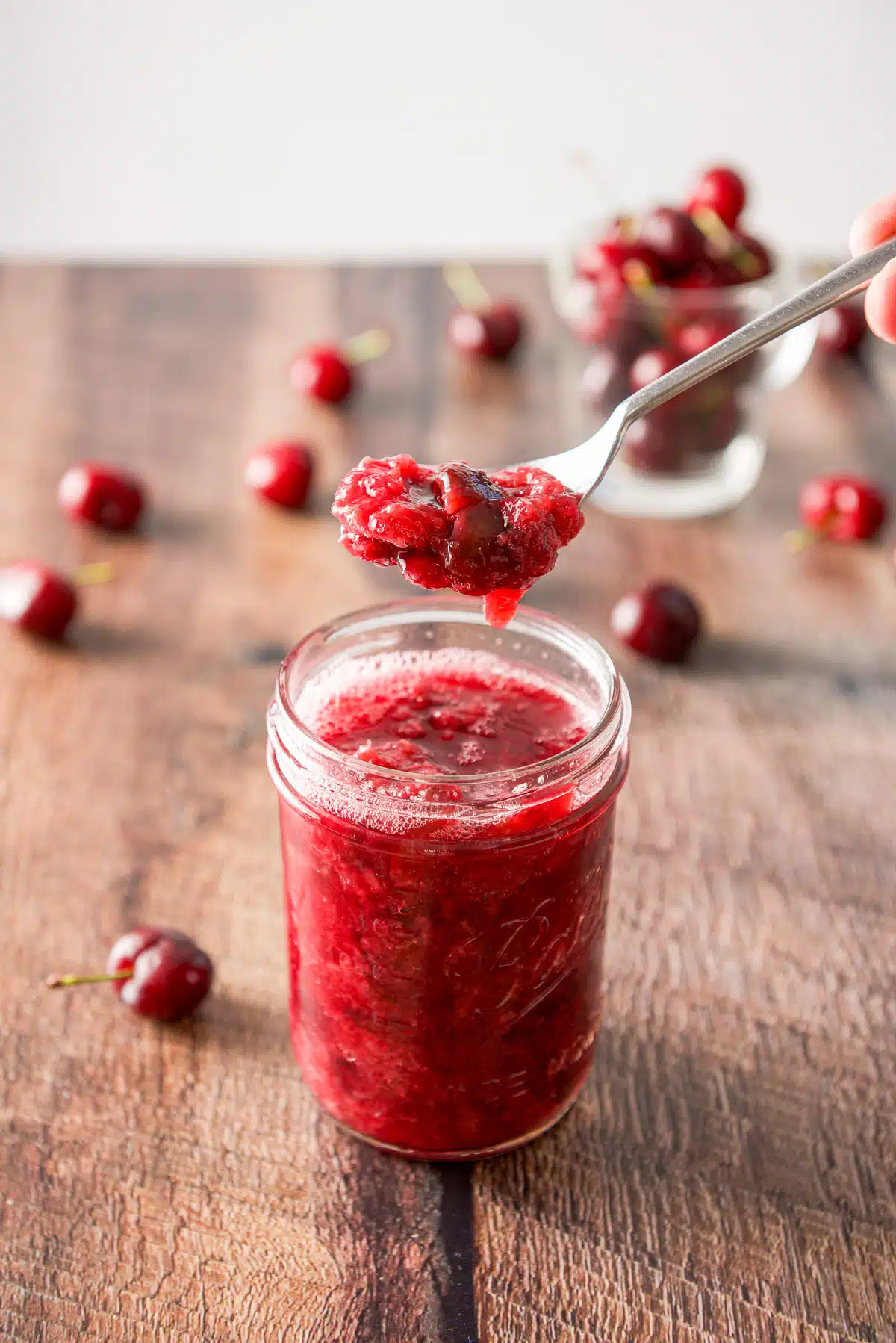 A spoonful of sauce held over a jar filled with it along with cherries in a glass and on the table