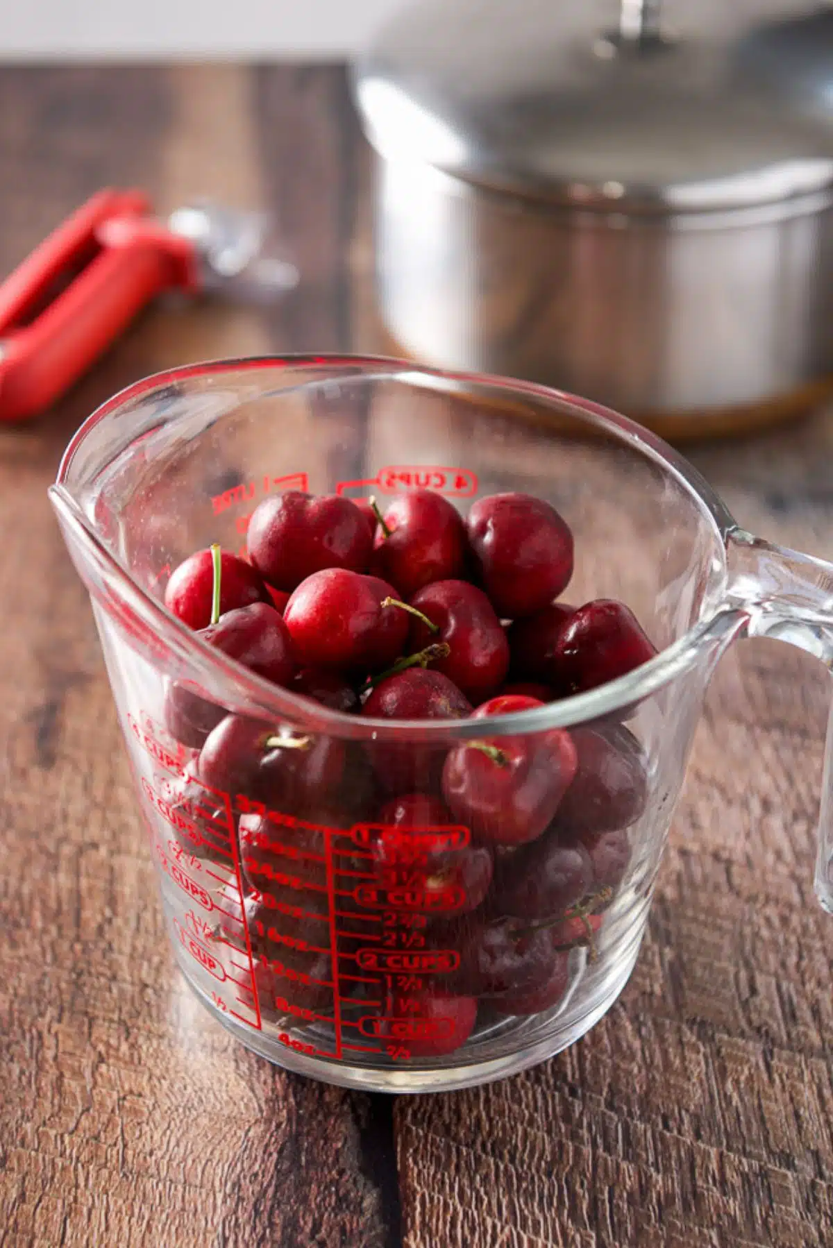 Cherries in a two cup measuring glass with a pitter and pan in the background