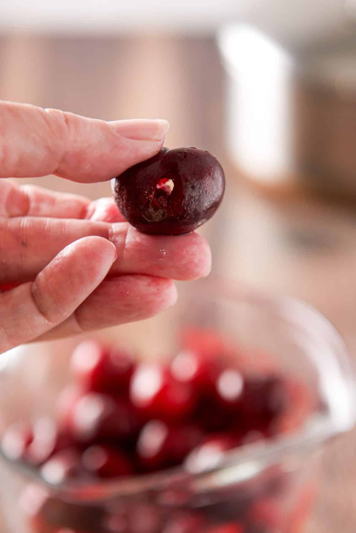 A female hand holding a pitted cherry with a hole in it over a jar of cherries