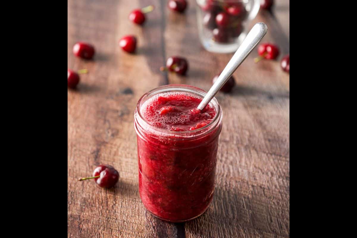 A jar of red fruit sauce with a spoon in it on a table with cherries