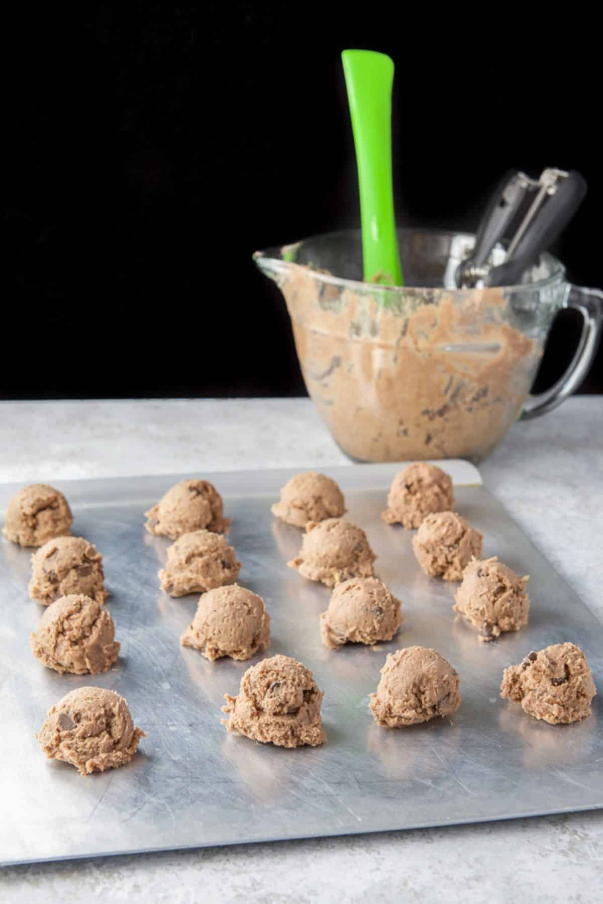 A cookie sheet with balls of chocolate cookie dough in front of a bowl of dough in the background