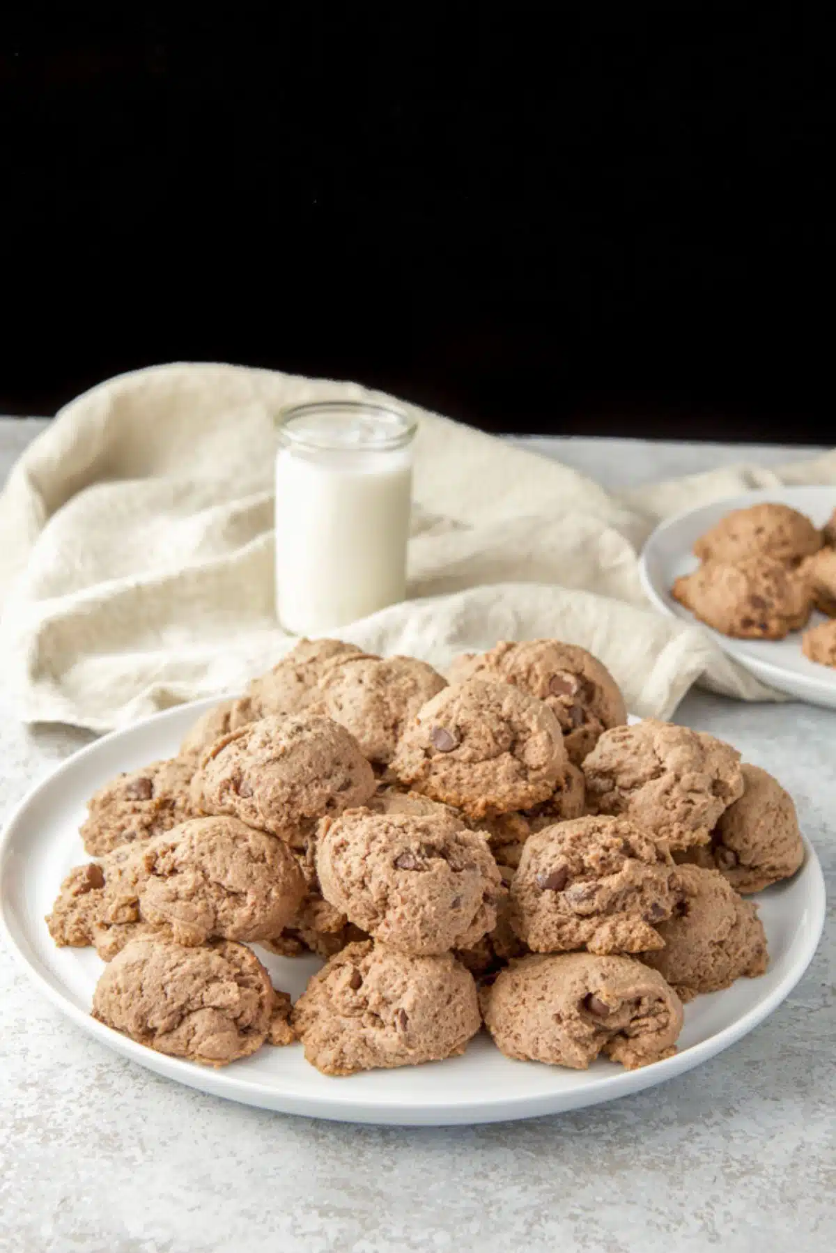 Pile of cookies on a big plate with a smaller plate of them in the background as well as a glass of milk