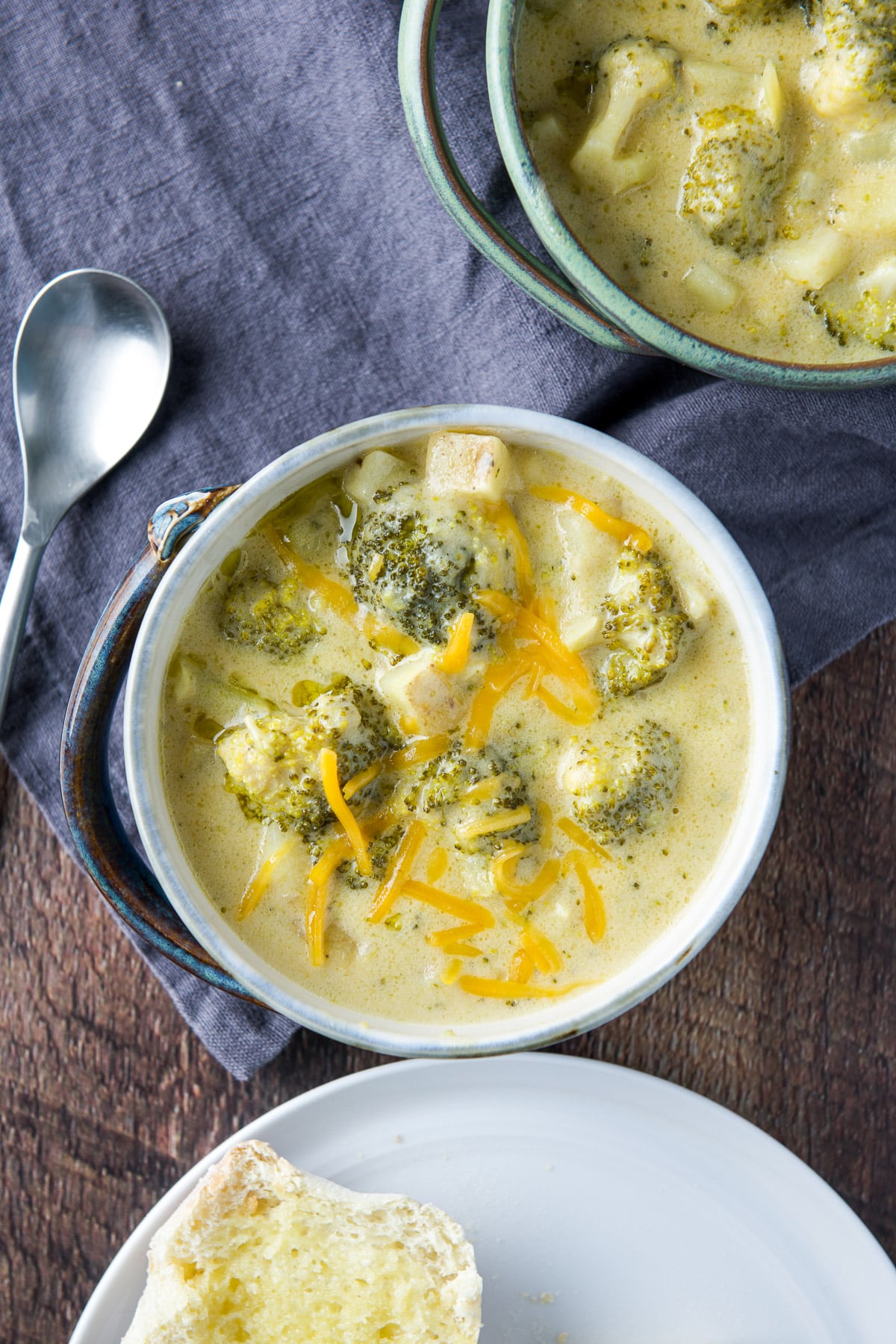 overhead view of two bowls of broccoli soup with a spoon and roll on a plate
