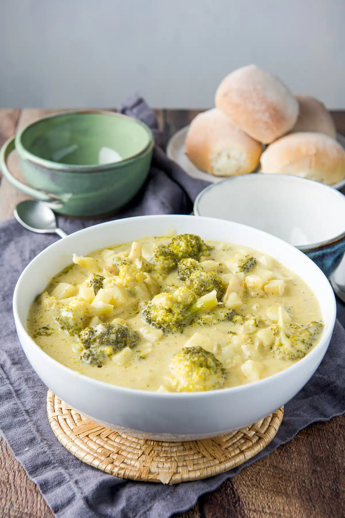 A large white bowl filled with the broccoli soup with two bowls in the back and a plate of rolls
