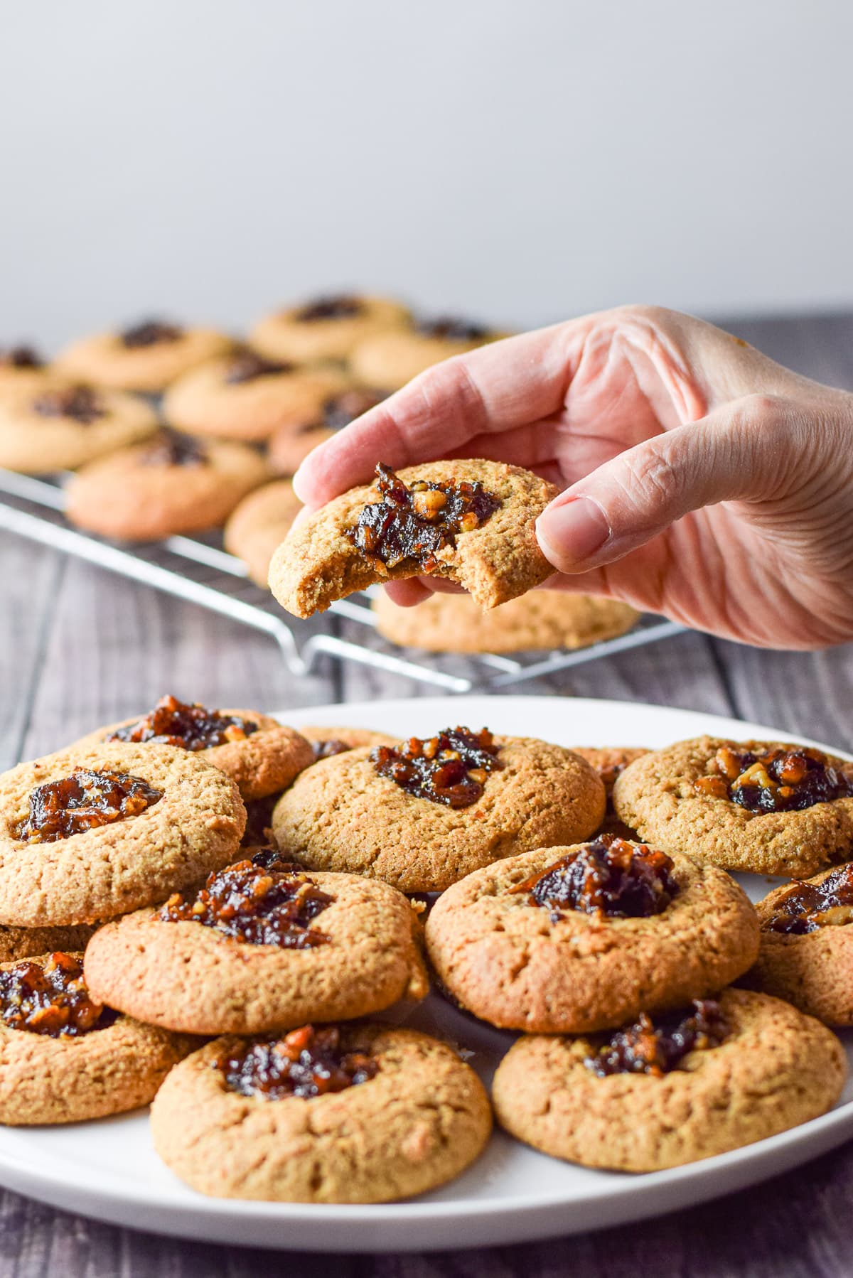 A female hand holding a cookie with a bite taken out it. There is a plate of cookies and a wire rack in the background