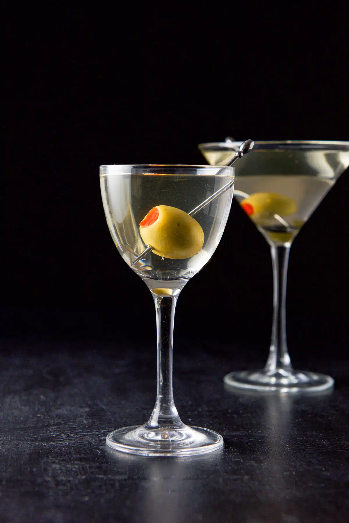 Vertical view of the smaller glass with the martini with a big olive in it. Regular martini glass in the background