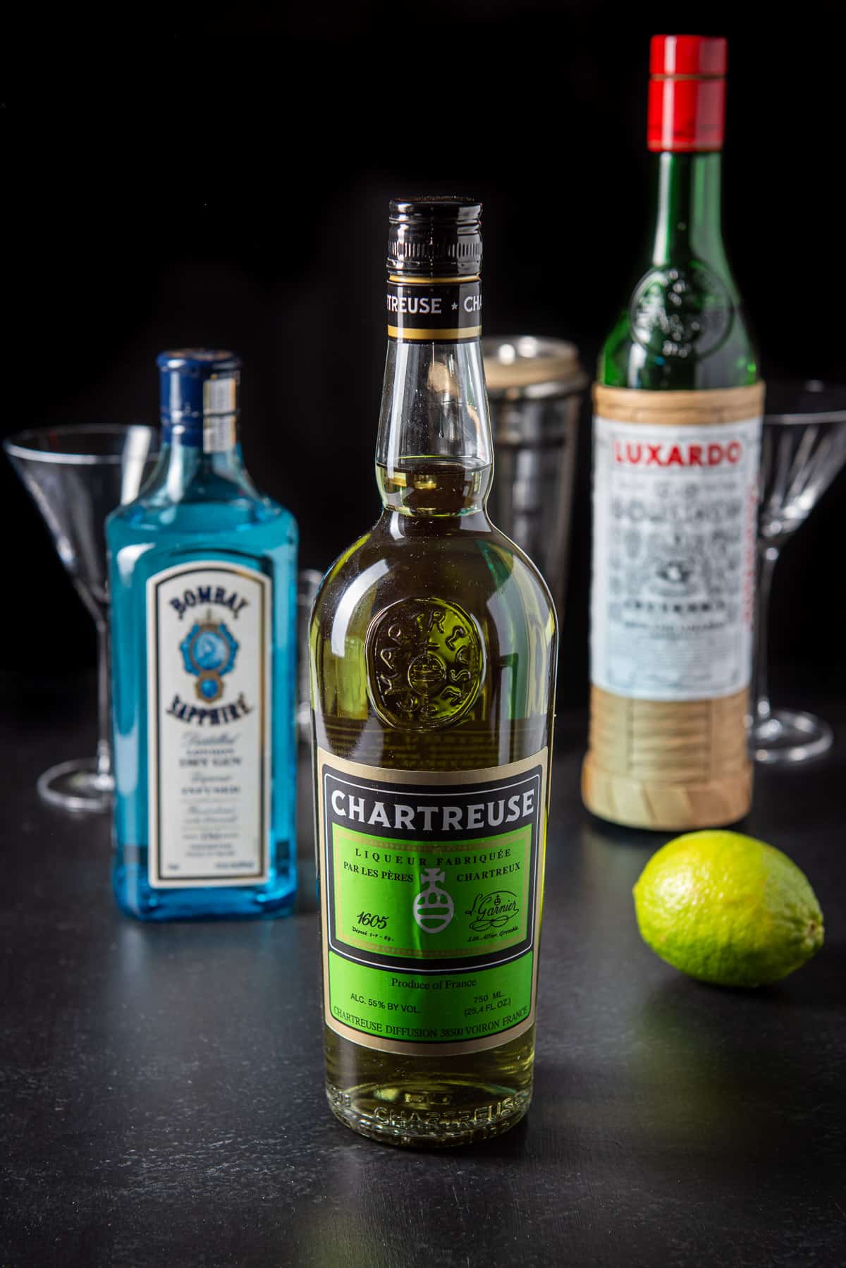 Bottles of Chartreuse, gin, maraschino cherry and a lime on a table with glasses and the shaker