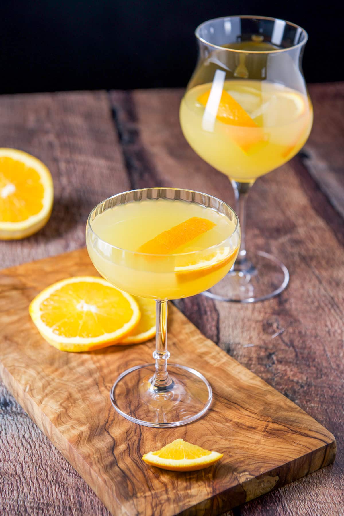 A coupe glass and tulip shaped glass with the orange cocktail in it with sliced oranges on the table