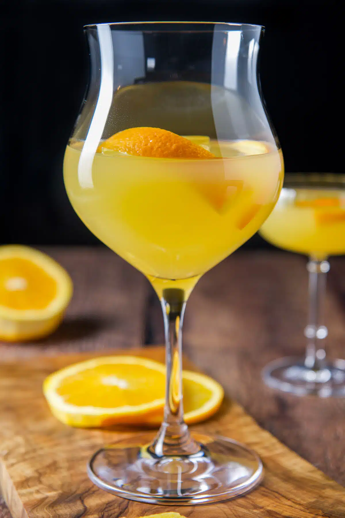 Vertical view of the tulip glass and coupe glass filled with the orange drink with more oranges on the table