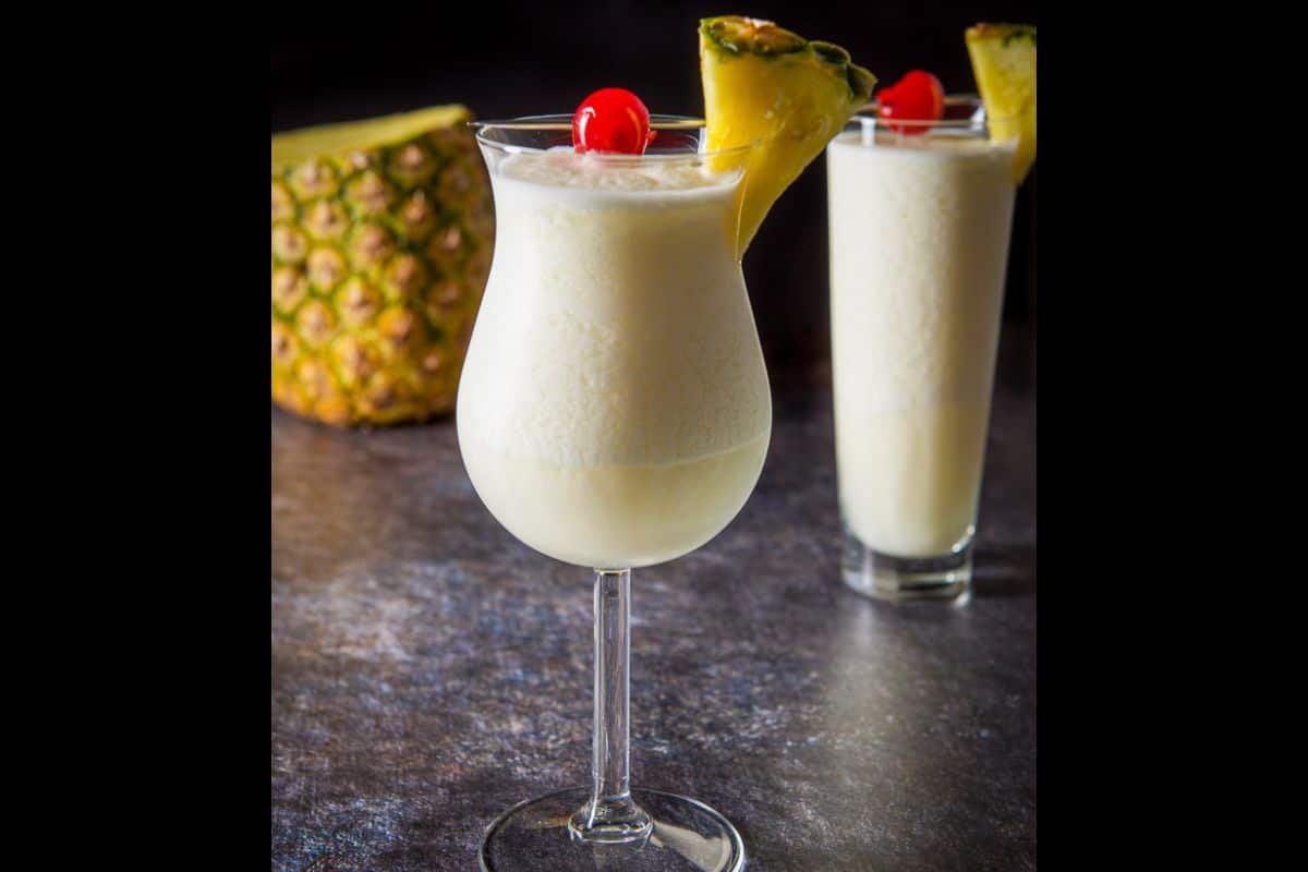 vertical view of the pina colada cocktail in two glasses with pineapple and cherries on the rim, and more pineapple in the back