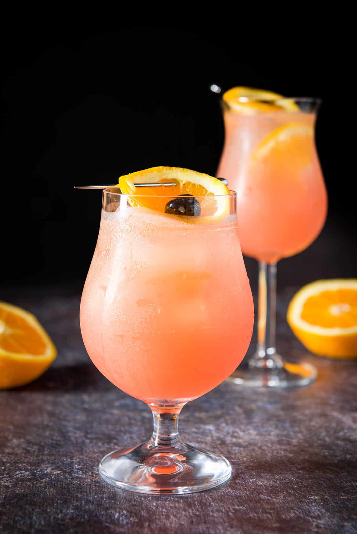 Short tulip glass with the orange cocktail with the orange and cherry as garnish. There is a tall glass in the background