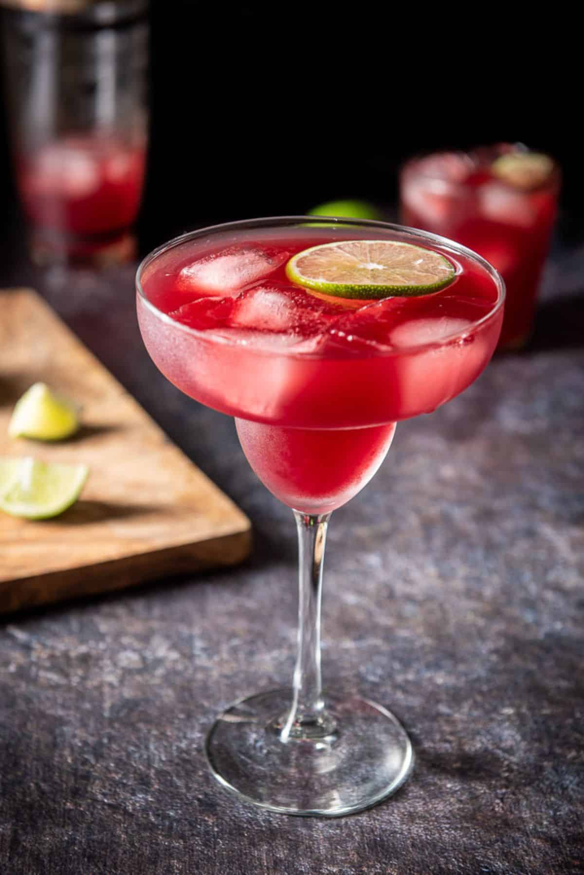 Classic margarita glass in front with liquid in it along with a floating lime wheel and ice
