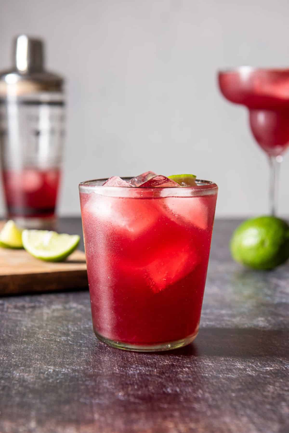 Double old fashioned glass filled with the red margarita with another glass in the back as well as limes and the shaker