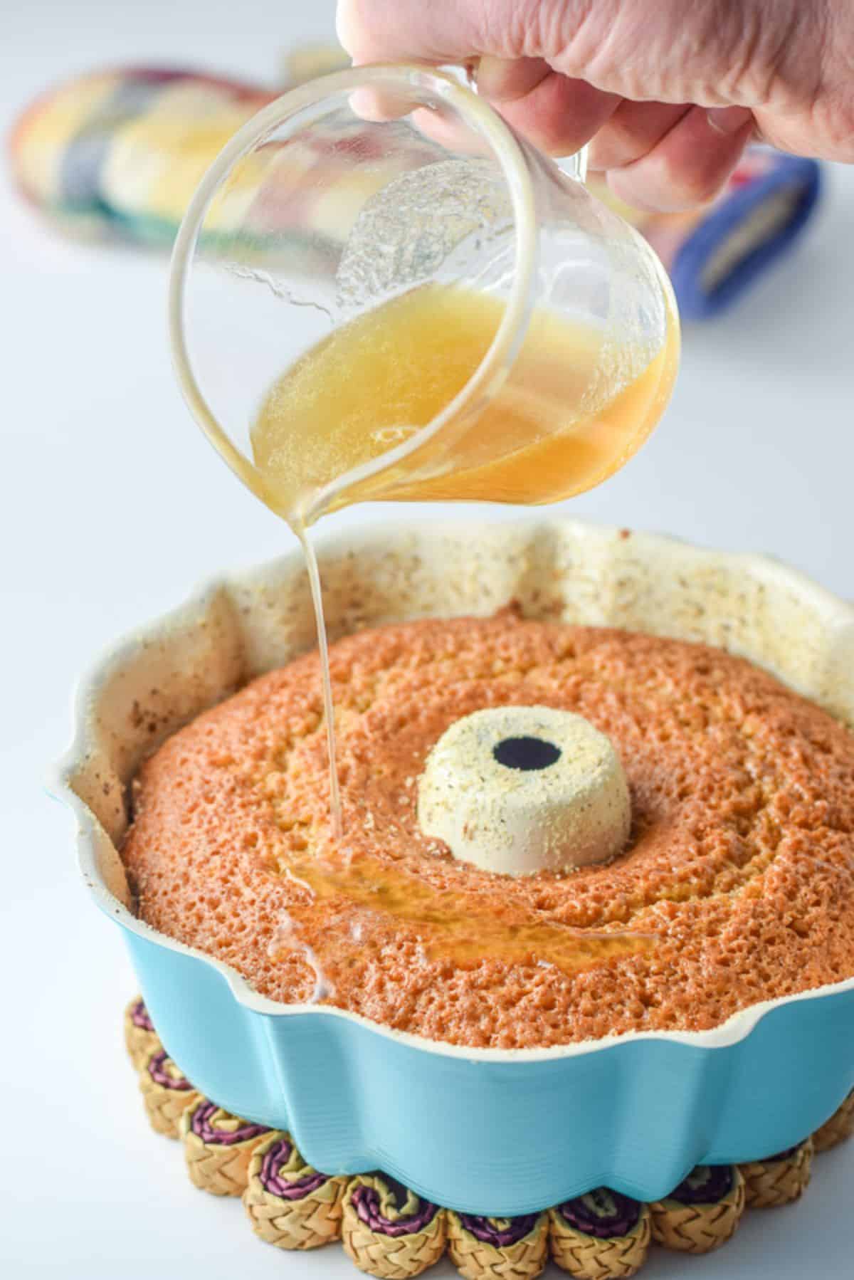 a hand holding a pitcher filled with a sauce being poured over the rum cake in the pan