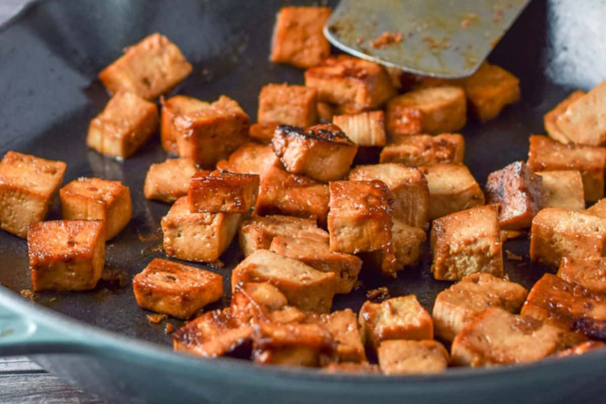 A blue cast iron pan with tofu that has been saut&eacute;ed. There are some noodles and a salad in the background